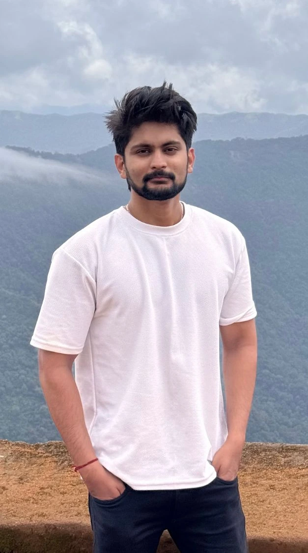 Amar Karthik standing behind a scenic view in a casual white shirt.